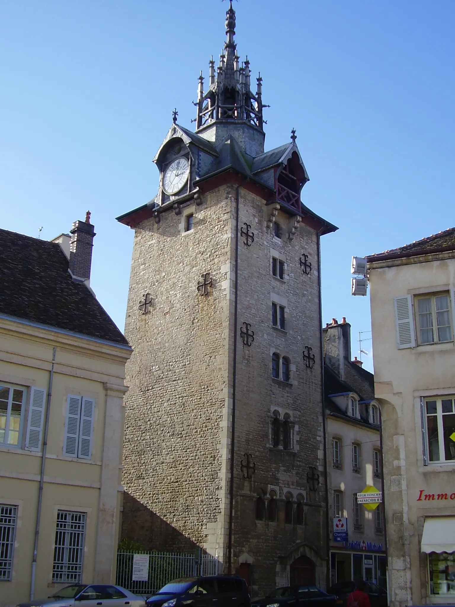 The stone clock tower of Beaune with its clock face, slate roof and pointed bell turret against a blue sky